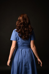 3/4 portrait of brunette lady wearing blue dress, facing away from camera. posed on black studio...