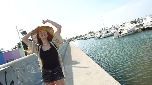 Young attractive smiling woman tourist walking, dancing and enjoying her life and weather on sunny summer day in ea port. Wearing stylish clothes, shorts and straw hat. Travel and journey concept.