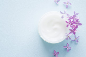Fototapeta na wymiar Opened white jar of herbal night cream for women on pastel blue table. Beautiful branch of purple lilac. Care about clean and soft face, hands, legs and body skin. Fresh flowers. Empty place for text.