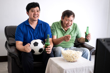 Young Asian Man and father watching soccer match on tv and cheering football team, celebrating with beer and popcorn at home, sports and entertainment concept