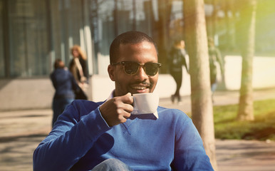 Portrait of smiling confident young African-American businessman in formal wear sitting at bench in city park and drinking hot coffee.Blurred background.Horizontal