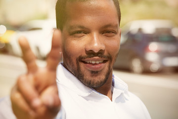 Closeup portrait of confident young African-American man hipster in white shirt showing fingers symbol V.Blurred streets on background.