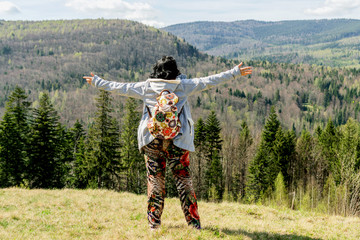 Adult traveling woman, with backpack, stands on top of a mountain peak, with her hands raised up, feeling free and success.