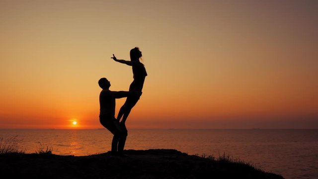 Amazing sillhouettes of beautiful couple doing acro-yoga on the sea or ocean beach at sunrise background, slow motion. Romantic scene. Healthy lifestyle, sport concept.