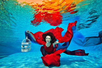 A young girl swims underwater at the bottom of the pool, plays with red and blue fabric, holding a...