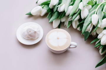 Fototapeta na wymiar Delicious fresh morning cappuccino coffee with flower drawn on its milk foam, marshmallows on the side and a big bunch of white tulips on the pastel pink table background