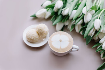 Delicious fresh morning cappuccino coffee with flower drawn on its milk foam, white chocolate cookies on the side and a big bunch of white tulips on the pastel pink table background