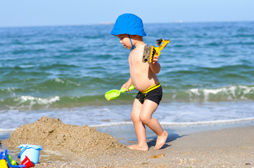 little cute baby boy on the beach playing with a yellow excavator in a blue panama