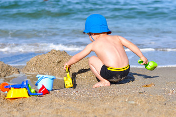 little cute baby boy on the beach playing with a yellow excavator in a blue panama