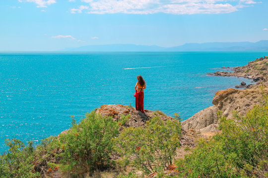 A woman is standing alone and on the edge of a cliff and looking over the blue Sea