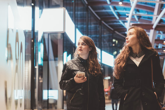 Two young beautiful caucasian women friends outdoor in the city night using smart phone hand hold strolling - technology, social network communication concept