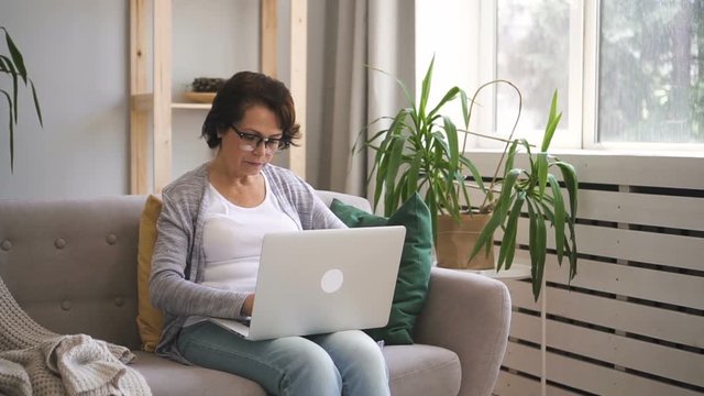 Elderly woman is using laptop sitting on sofa at home, seniora wearing glasses is typing, looking at pc screen, having good time near window with green plant in modern interior. Concept: mature person