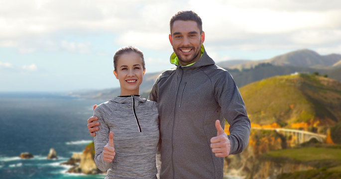 fitness, sport and gesture concept - smiling couple outdoors showing thumbs up over bixby creek bridge on big sur coast of california background