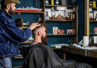 Man with beard and mustache in hairdressers chair in front of mirror, shelves on background. Barber styling hair of bearded client with wax by hands. Barbershop concept. Hipster client got hairstyle