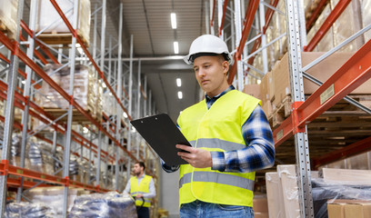 logistic business, shipment and people concept - male worker or supervisor with clipboard in reflective safety vest at warehouse