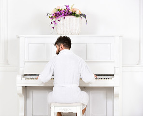 Man sleepy in bathrobe sit in front of piano musical instrument in white interior on background, rear view. Talented musician concept. Man in bathrobe enjoys morning while playing piano.