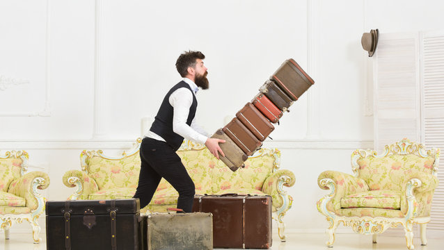 Baggage insurance concept. Man with beard and mustache in classic suit delivers luggage, luxury white interior background. Porter, butler accidentally stumbled, dropping pile of vintage suitcases.