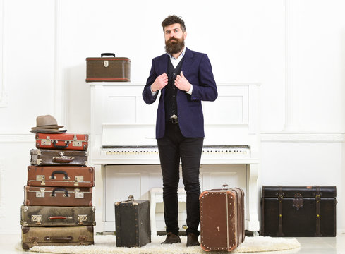 Man, traveller with beard and mustache with luggage, luxury white interior background. Macho elegant on strict face stands near pile of vintage suitcase, ready for vacation. Luggage and travel concept