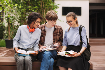 Fototapeta premium Group of smiling students sitting on bench and reading books in courtyard of university. Company of young students study together outdoors