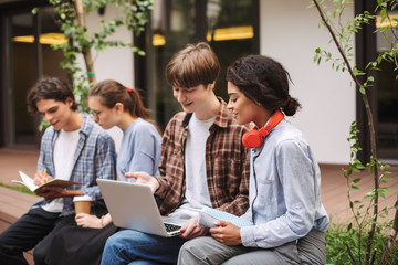 Smiling boy and girl sitting on bench and using laptop while spending time in courtyard of...