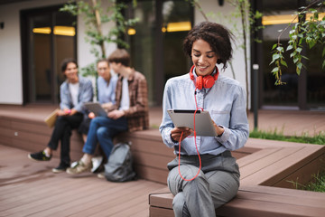 Smiling lady sitting on bench in courtyard of university with tablet in hands and red headphones. Young woman sitting and using tablet with students on background