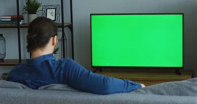 View From Behind Of The Man In The Jeans Blue Shirt Sitting On The Couch In The Living Room And Watching TV With Green Screen. Chroma Key. Indoors