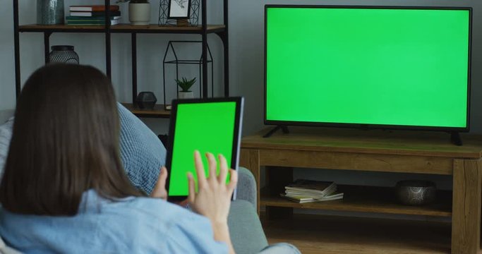 View From Behind On The Young Woman With Dark Hair Sitting At Home, Watching TV With Green Screen And Scrolling On The Green Screen Of The Black Tablet Computer. TV In Focus. Chroma Key. Indoor