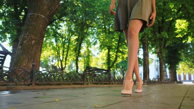 Sexy legs with high beige heels walking in park of city. Woman walking on boulvard alone. Attractive girl in short dress at summer season. Slow motion.