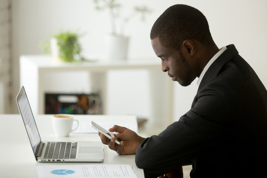 Focused serious young African American person checking email on smartphone, looking at phone while working at laptop in office, reading news online at cell, browsing web and chatting in social media