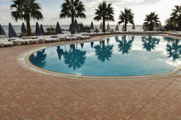 Empty swimming pool during a bit cloudy day during summer vacation. View of blue water in pool, sunbeds, umbrellas, palms, horizon and sea in the background. Luxury place for relax in paradise.