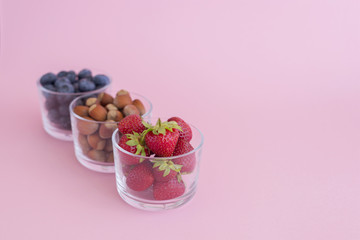 Three glass cups with strawberry, blueberry and hazelnuts on pink background