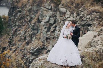 A fashionable bridegroom in a black suit and a beautiful bride in a lace dress with a long veil embrace against the background of rocks and stones. 