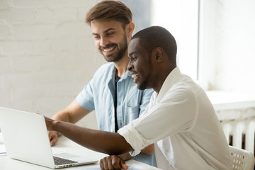 Obraz premium Multiethnic office workers laughing watching funny videos at laptop, African American guy showing something to Caucasian colleague. Diverse team at casual break, smiling, having fun, good relations