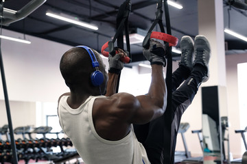 A black muscular guy in a white T-shirt and headphones hangs on the gym rings and understands his legs. Close photo.