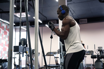 A black muscular man in a T-shirt and headphones shakes his hands with a simulator in the gym.