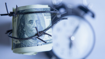 US Dollar money wrapped in barbed wire against clock background as a symbol of economic warfare, sanctions and embargo busting
