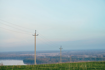 Atmospheric landscape with power lines in green field on background of river under blue sky. Electric pillars with copy space. Wires of high voltage above ground. Electricity industry.