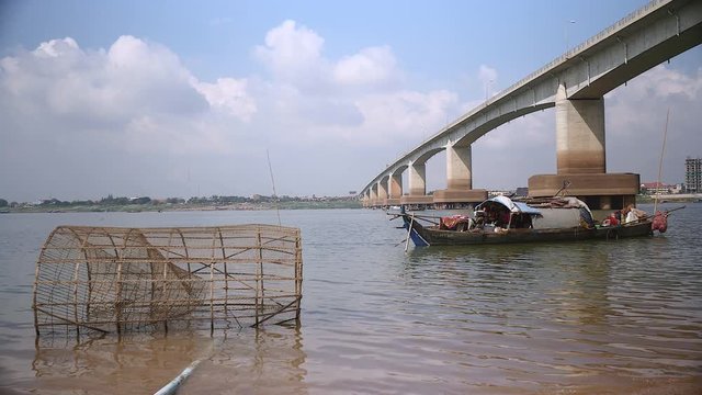 Small wooden fishing boat on the river nearby a bridge; Traditional bamboo fish trap in the foreground