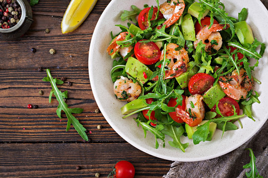 Fresh salad bowl with shrimp, tomato, avocado and arugula on wooden background close up. Healthy food. Clean eating. Top view. Flat lay.