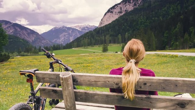 Girl sits on bench in front of beautiful landscape from forest, field and mountains. Rest during bike trip in the Alps. Meditation in heavenly place. Cycling in the mountains.