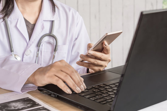 doctor sitting at desk working on computer laptop and hand holding mobile phone checking information of patient 