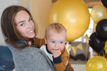 Beautiful young woman with cute baby smiling and looking at camera while standing in room full of party balloons