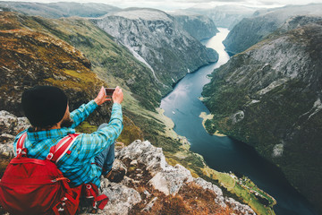Naklejka premium Man backpacker taking photo by smartphone relaxing on mountain cliff traveling in Norway adventure lifestyle active vacations modern technology connection concept