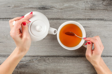 Table top view on young woman hands holding white ceramic teapot in one and silver spoon above cup...