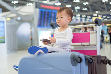 Little traveler, Cute Asian 18 months / 1 year old toddler boy child holding passport with suitcase, sitting on trolley at airport, waiting for departure, Family travel & vacation with kid concept