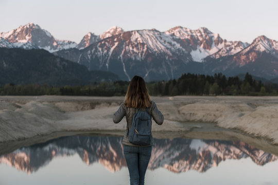 Young adult woman standing in front of a lake with glowing mountains reflected in it at sunrise