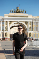 Fototapeta premium Young stylish man tourist walking near General Staff Builsing arch in the palace square in St. Petersburg. He wearing in black jeans, black T-shirt, black cap with riund sunglasses