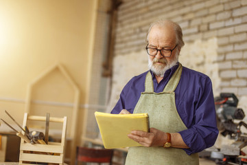 closeup photo of handsome old craftsman writing something in the notepad while working indoors