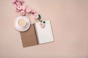 White cup with cappuccino, sakura flowers, notebook on a pastel pink background.