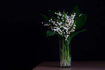 Flowers of lilies of valley on black background in a glass vase. Photo with copy blank space.